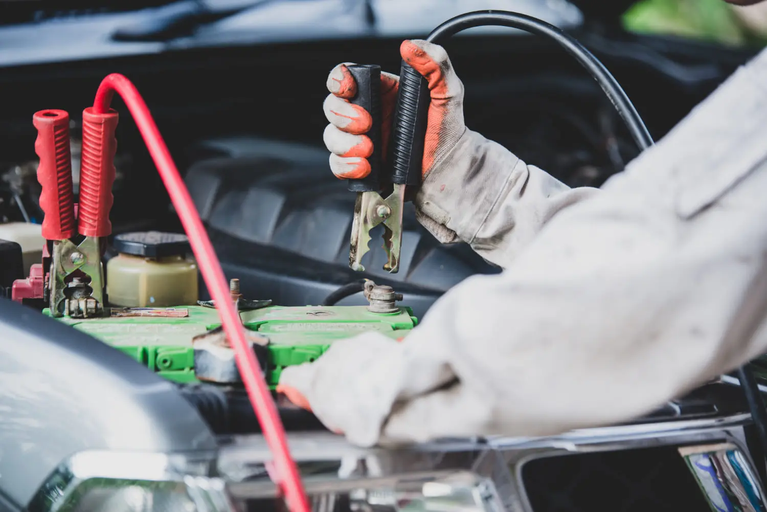 Gloved mechanic connecting black jumper cable clamp to car battery terminal under hood